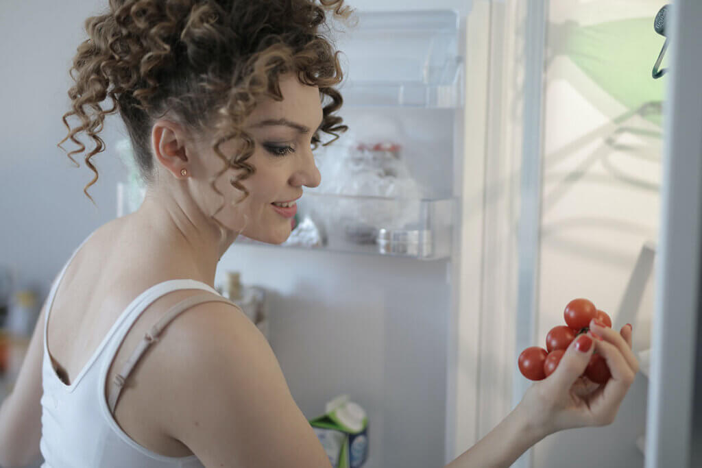Frau mit Locken steht vorm geoeffnetem Kuehlschrank und haelt Tomaten in der Hand.