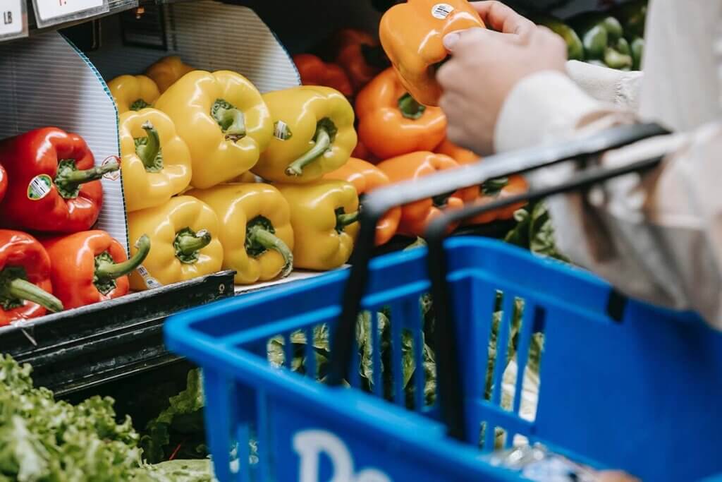 Person mit blauem Einkaufskorb am Arm steht vor einem Gemueseregal und haelt eine orangene Paprika in der Hand.