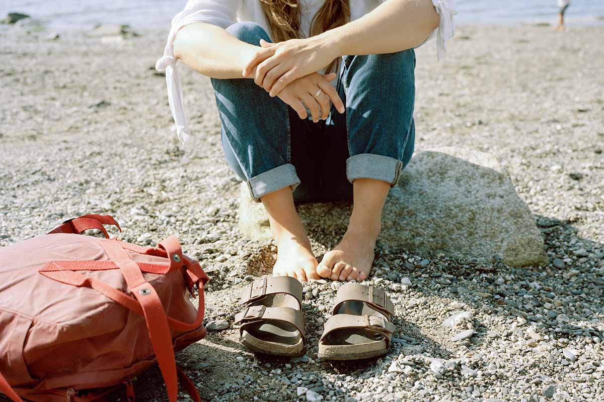 Person mit Sandalen sitzt am Strand im Sand.