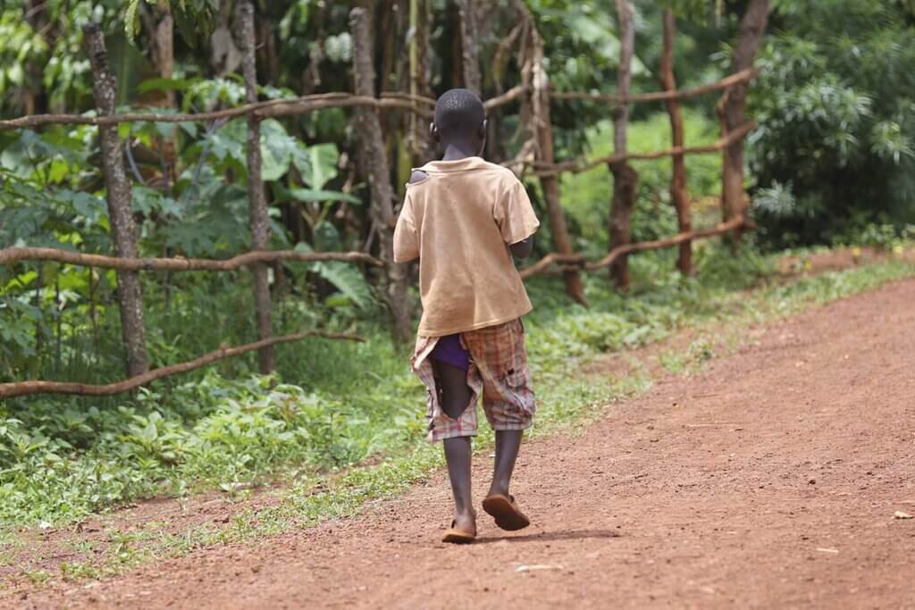 Ein Kind laeuft mit zerrissener Kleidung auf einer sandigen Strasse neben einem Wald.