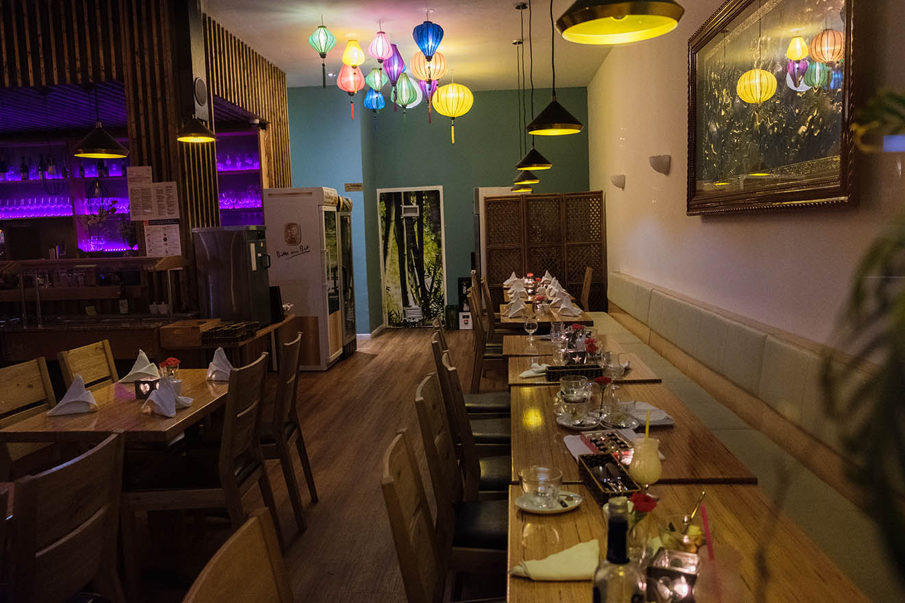 Ein Restaurant mit verschiedenen Lampen und unbesetzten Tischen. Die Bar ist lila beleuchtet.