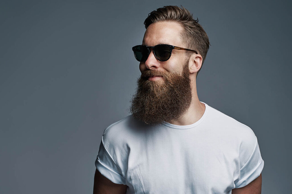 Mann mit Bart und Sonnenbrille steht vor einer grauen Leinwand.