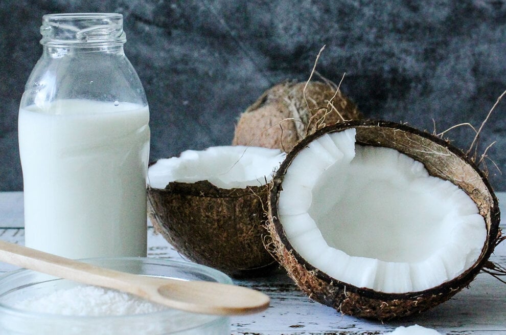 Offene Kokosnuss liegt neben einer Flasche mit Kokosmilch und einem Holzloeffel