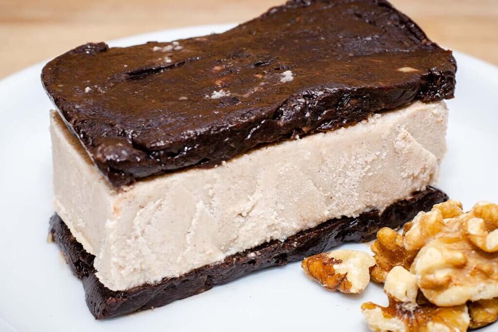 Vegane Eisschnitte mit braunem schokoladigem Boden und Topping, auf einem weissen Teller, garniert mit Walnuessen.