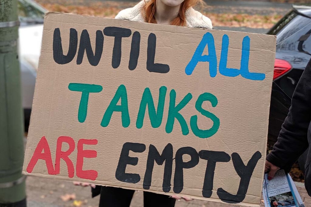 Person steht mit Pappschild auf der Strasse auf dem in bunten Buchstaben steht: Until all tanks are empty.