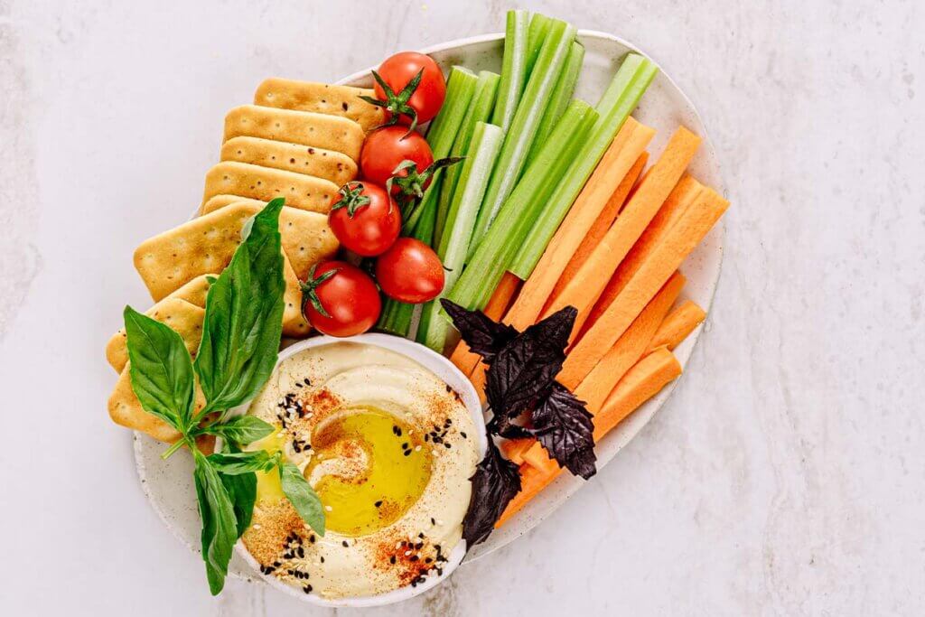 Teller mit Gemuesesticks von Karotte, Sellerie sowie Tomaten und Crackern, die neben einer Schale Hummus liegen.