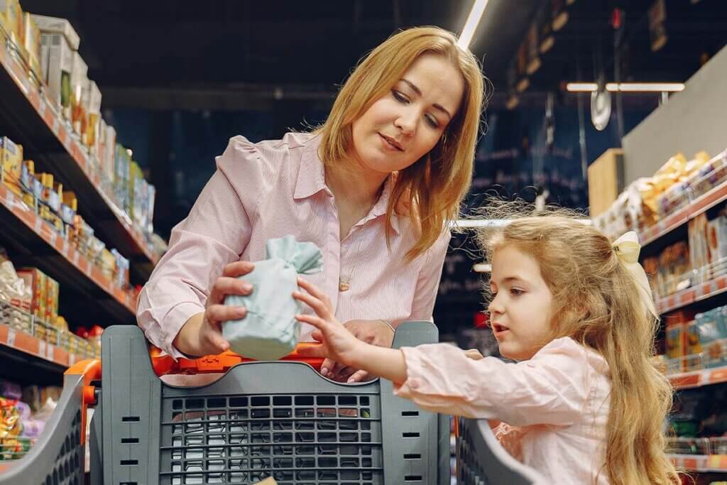 Blonde Frau mit blondem Kind in rosa Blusen stehen vor einem Einkaufswagen im Supermarkt und betrachten ein Produkt, dass die Frau in der Hand haelt.
