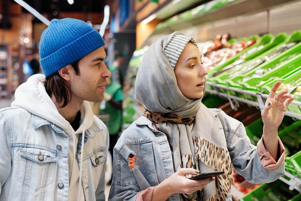 Mann mit blauer Muetze und Jeansjacke steht neben einer Frau mit grauer Muetze und Schal um den Kopf vor einem Supermarktregal.