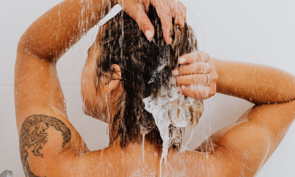 Person shampooniert sich ihr schwarzes Haar unter der laufenden Dusche.