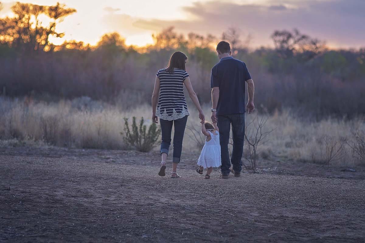 Mann und Frau halten ein kleines Kind in der Mitte an der Hand und laufen bei Sonnenuntergang auf einer Wiese.