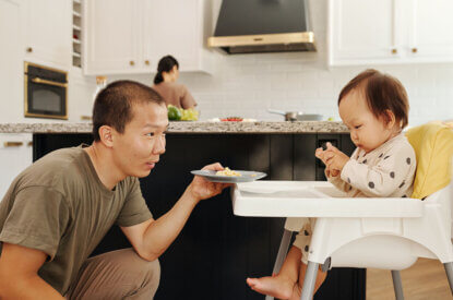 Kleikind sitzt im Stuhl in einer Küche während eine Frau im Hintergrund kocht und ein Mann dem Kind ein Teller mit Essen reicht.