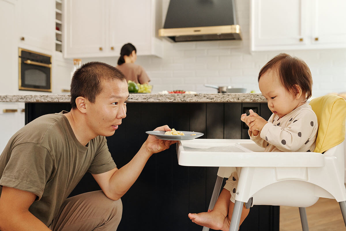 Kleikind sitzt im Stuhl in einer Küche während eine Frau im Hintergrund kocht und ein Mann dem Kind ein Teller mit Essen reicht.