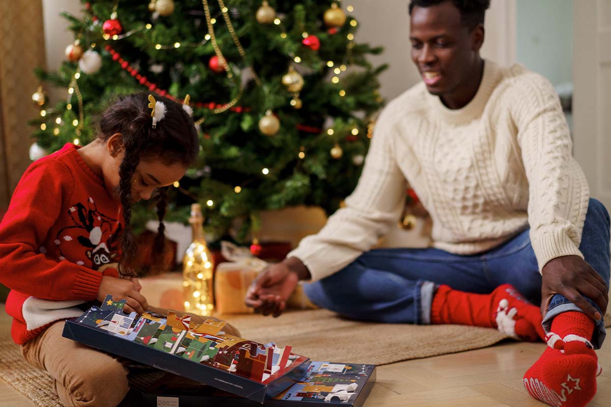 Ein Kind oeffnet einen Adventskalender auf dem Boden vor einem Weihnachtsbaum.