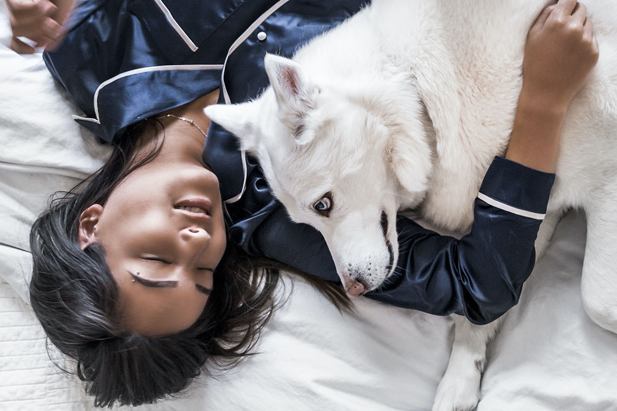Eine Frau in blauem Seidenpyjama liegt mit einem weissen Hund auf einem Bett.