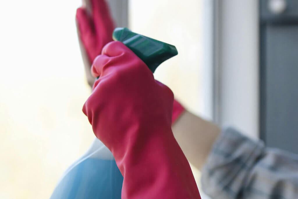 Eine Person putzt mit Handschuhen und einem Reiniger ein Fenster.
