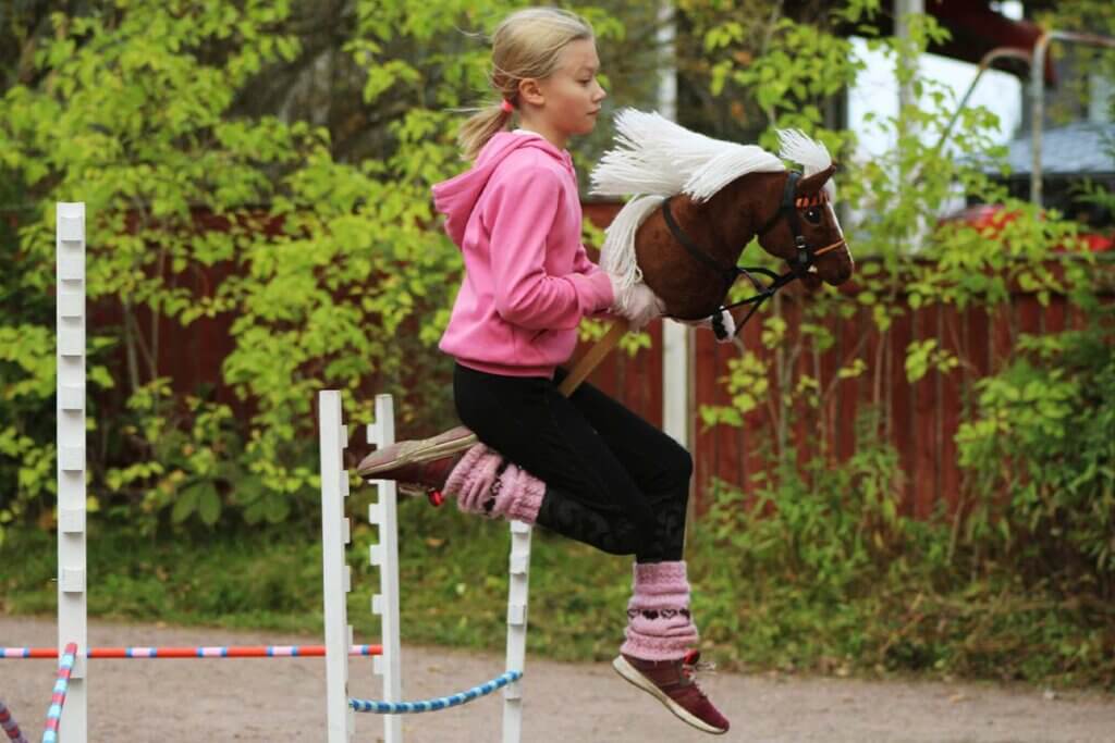 Ein Kind springt mit einem Steckenpferd ueber ein Hindernis.
