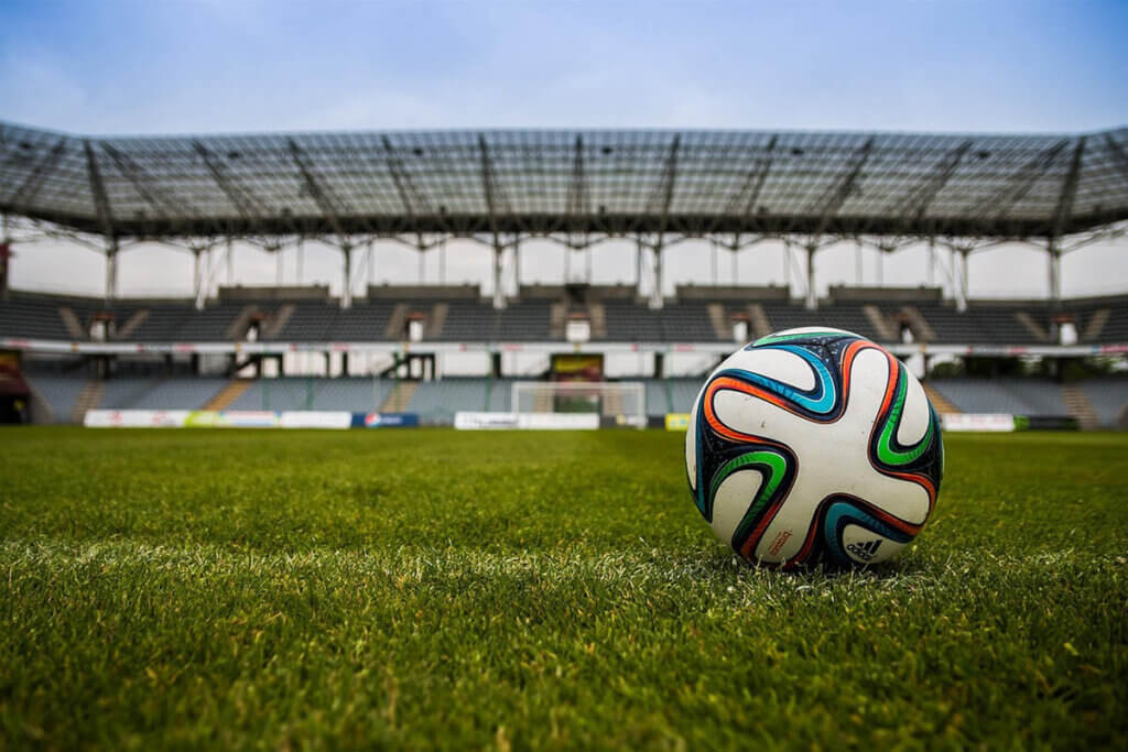 Ein Ball im Fokus in der Mitte eines leeren Fußballstadions mit grünem Rasen, der Ball befindet sich im rechten Bereich des Bildes.