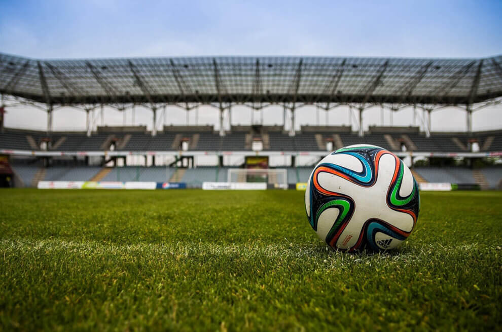 Ein Ball im Fokus in der Mitte eines leeren Fußballstadions mit grünem Rasen, der Ball befindet sich im rechten Bereich des Bildes.