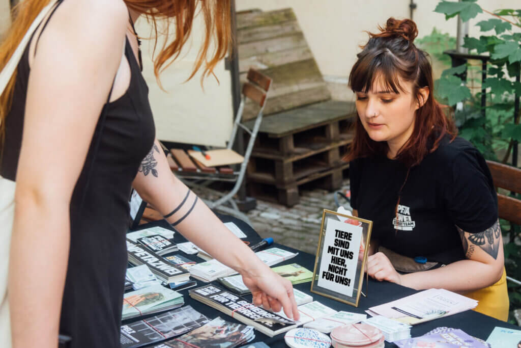 PETA Tabling. Eine junge Frau sitzt und berät, eine andere Frau steht davor und schaut sich Auslage an.
