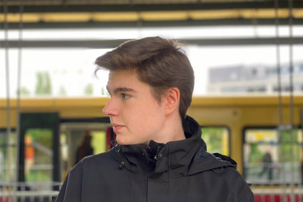 Milan steht an einem Bahngleis schaut von sich aus nach rechts und trägt dabei eine Kurzhaarfrisur und eine schwarze Windbreaker-Jacke.