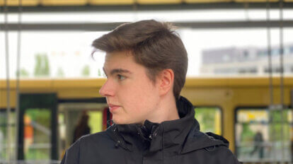 Milan steht an einem Bahngleis schaut von sich aus nach rechts und trägt dabei eine Kurzhaarfrisur und eine schwarze Windbreaker-Jacke.